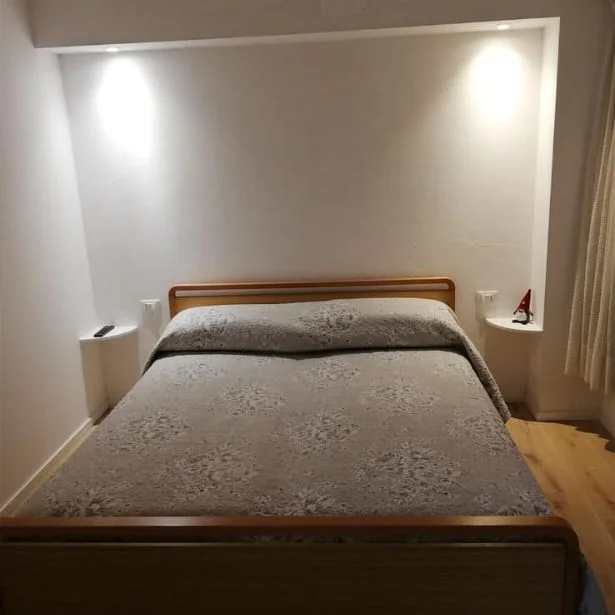 A double bed fills a softly lit bedroom with recessed wall lighting and pale curtains.